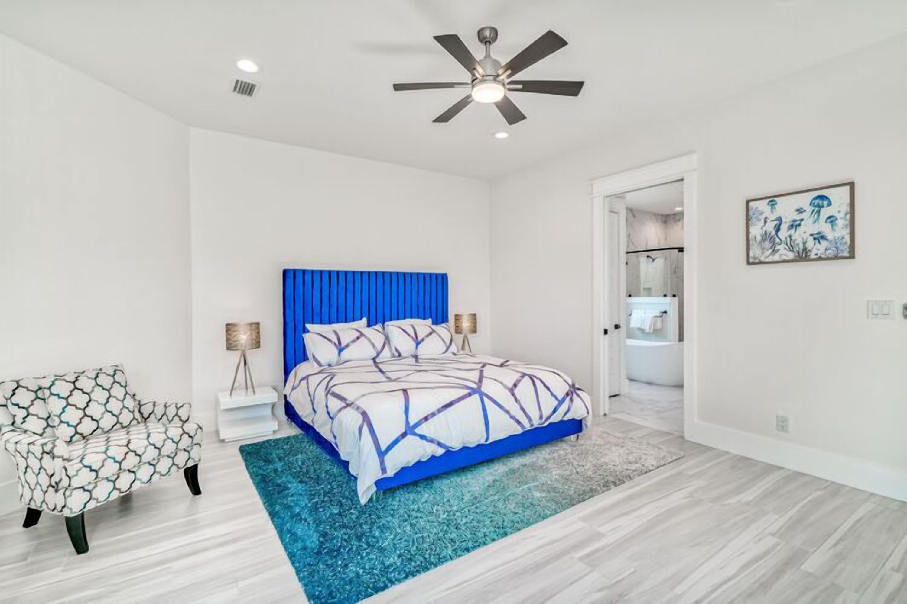 Une chambre à coucher comprenant un lit, une chaise, un ventilateur de plafond et une porte donnant sur une salle de bain.