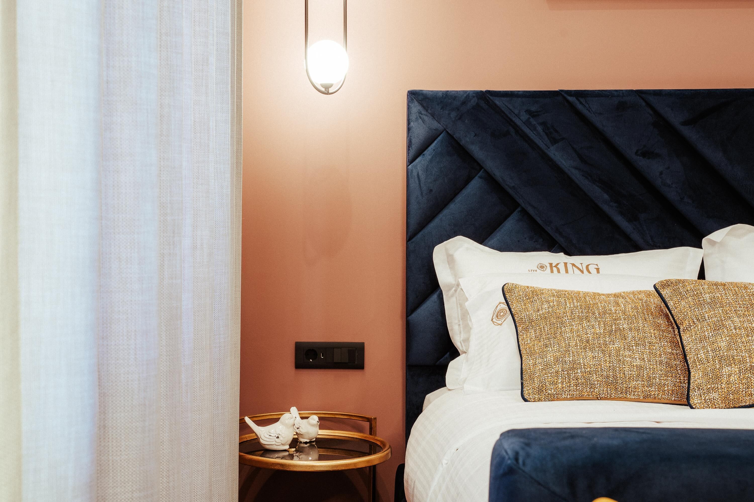 Habitación de hotel con una cama grande, una mesita con dos aves blancas, una lámpara de pared y un interruptor.