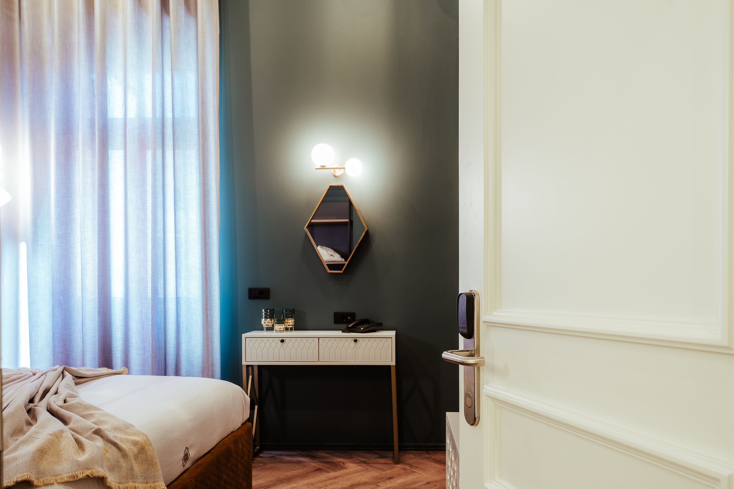 Hotellihuone, jossa on sänky, peilillä varustettu työpöytä, seinään kiinnitetty valaisin ja ovi.