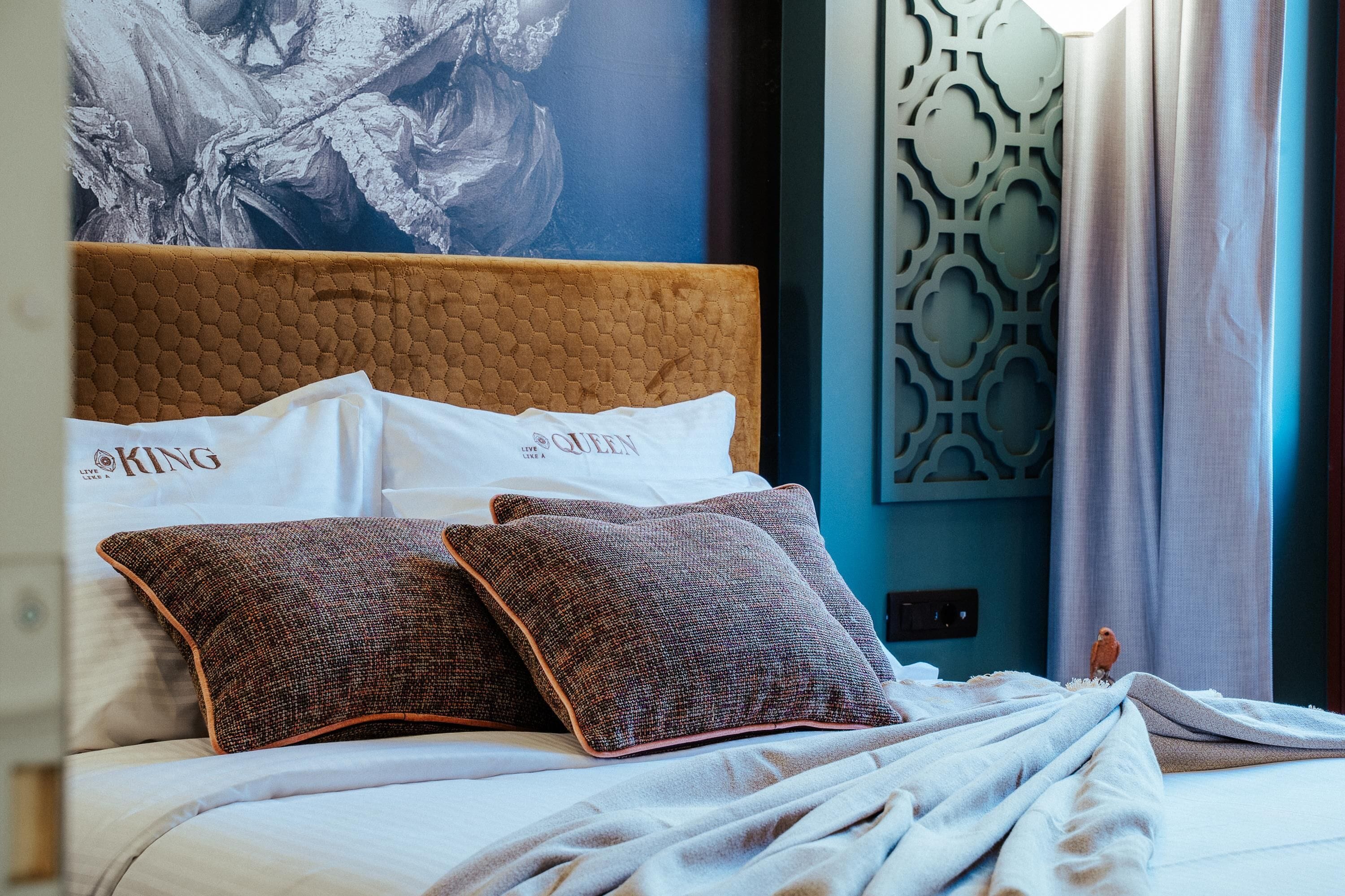 A neatly made bed with a brown headboard and two patterned pillows, a large abstract painting on the wall, and a decorative wall panel.