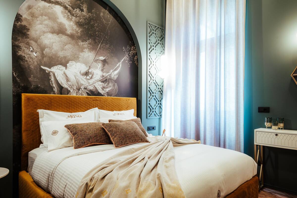 A bedroom with a large bed, a headboard with a mural, a nightstand, and a window with curtains.