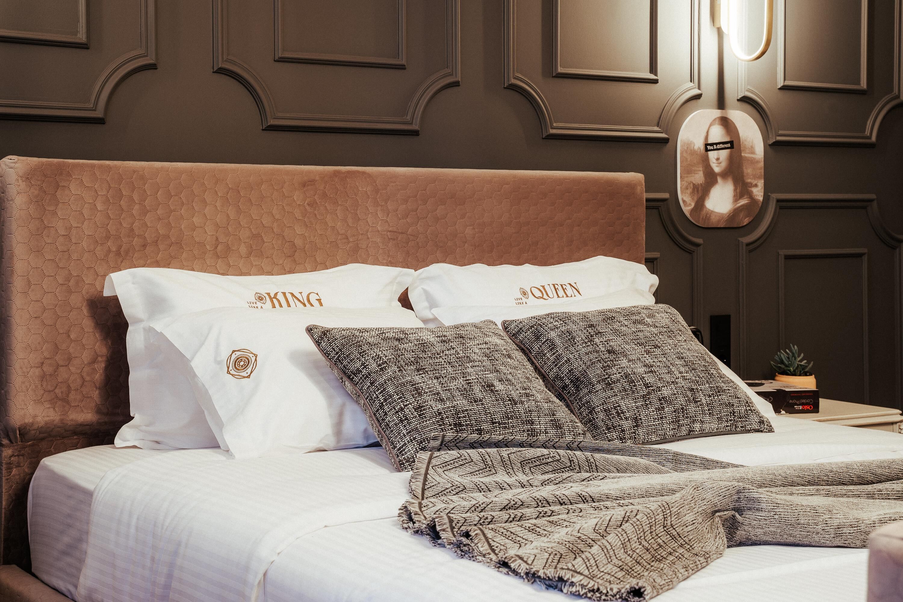 A neatly made bed with a brown headboard, two pillows with "KING" and "QUEEN" embroidered on them, and a patterned throw blanket.