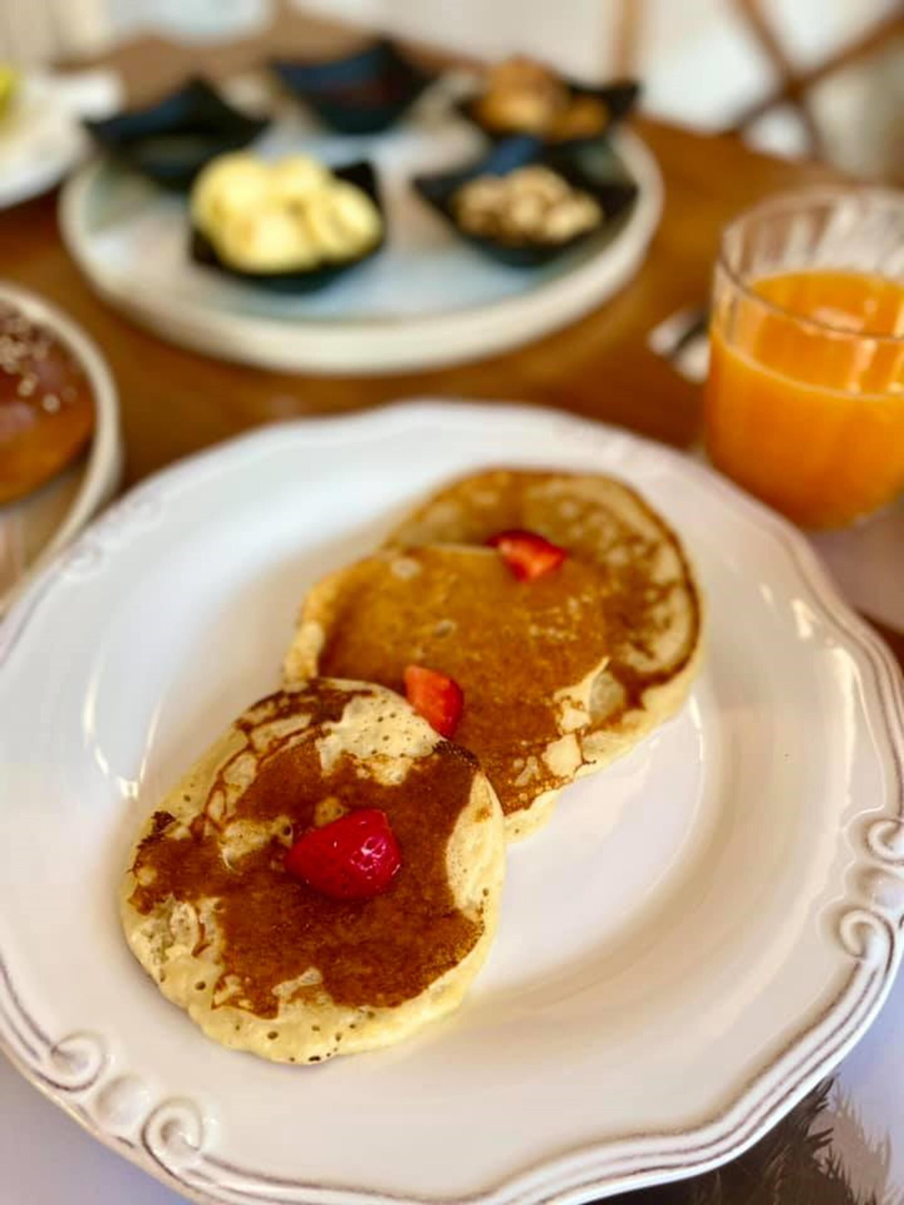 A plate with two pancakes topped with strawberries and syrup, a glass of orange juice, and a plate with various pastries in the background.