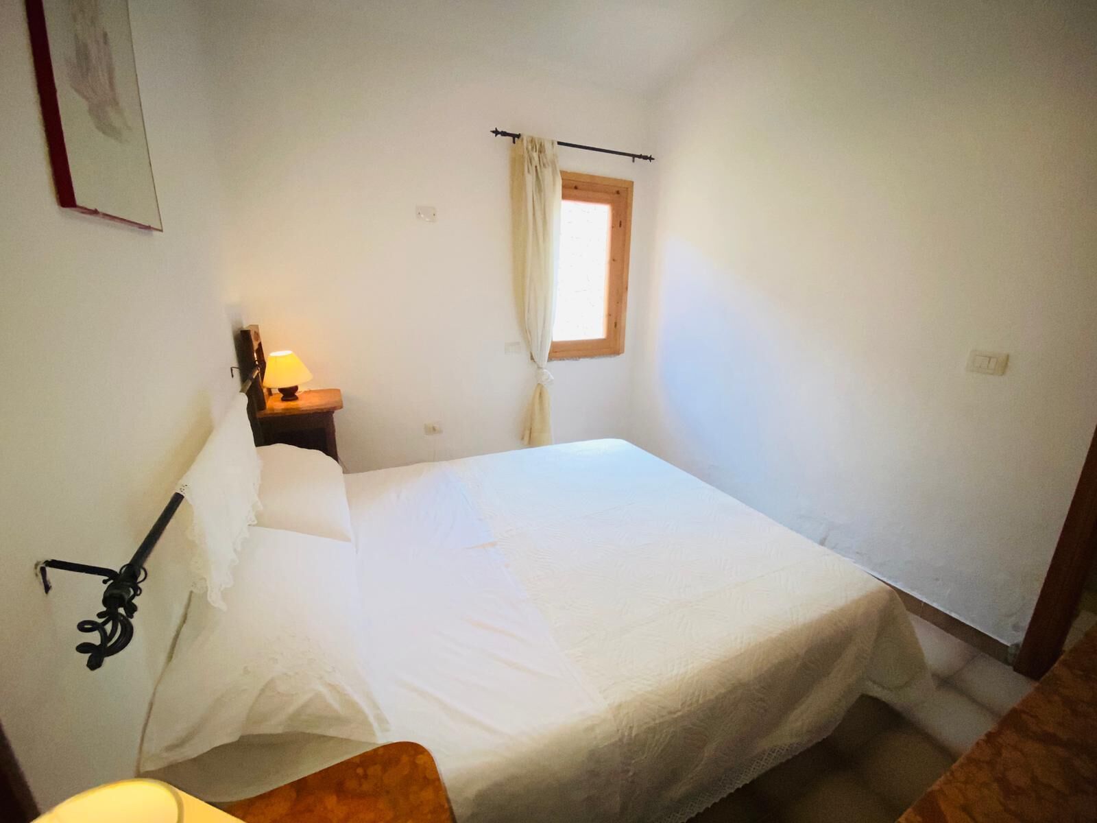Een kleine, eenvoudige kamer met een bed, een nachtkastje, een raam met gordijnen en een wandlamp.