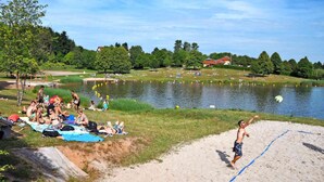 Ein Strand am See mit Menschen, die entspannen und Volleyball spielen, umgeben von viel Grün und Häusern.