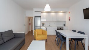 Un salon moderne comprenant un canapé, un fauteuil jaune, une table à manger avec des chaises et un coin cuisine avec des meubles blancs et des appareils électroménagers.