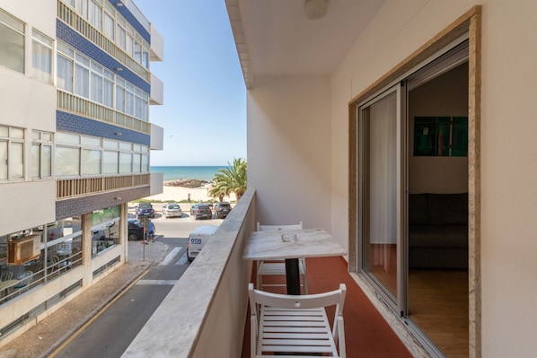 A balcony with a view of the beach, a table, and chairs.