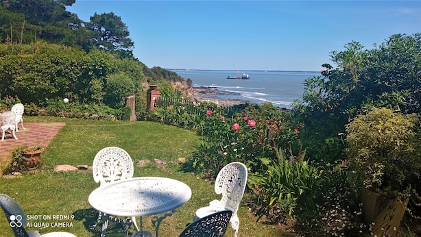 A garden with white metal chairs and a table, overlooking a coastal area with a view of the sea and a distant ship. - "Un Petit Coin de Paradis" (Saint-Nazaire)