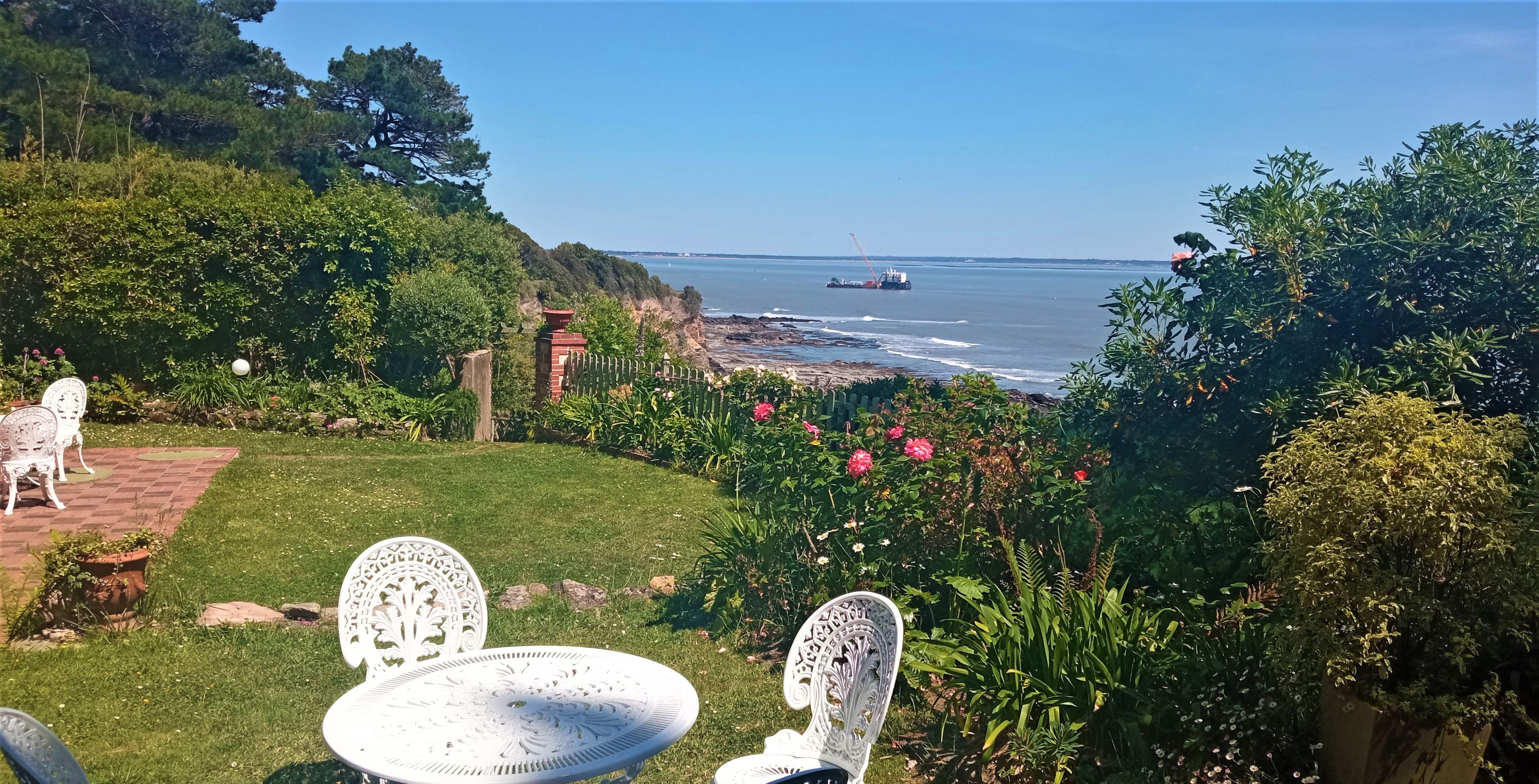 A garden with white metal chairs and a table, overlooking a coastal area with a view of the sea and a distant ship.