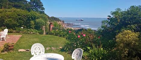 A garden with white metal chairs and a table, overlooking a coastal area with a view of the sea and a distant ship.