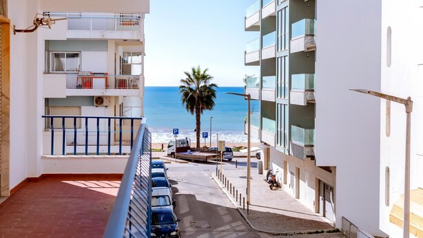 Vista desde el balcón de un apartamento con vista a una playa, con una fila de coches aparcados y una palmera al fondo.
