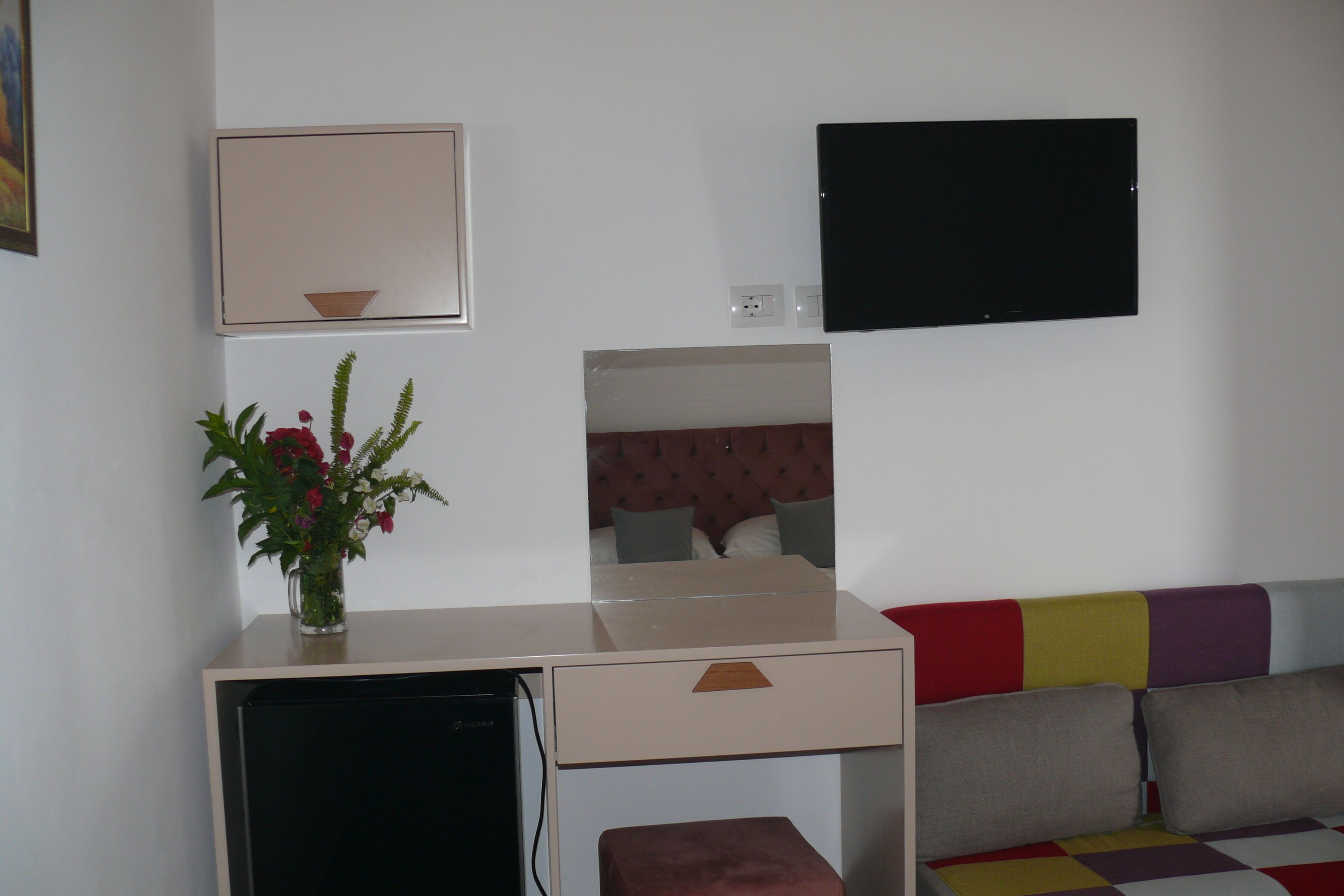 Una habitación con escritorio, refrigerador pequeño, espejo, televisor y sofá colorido.
