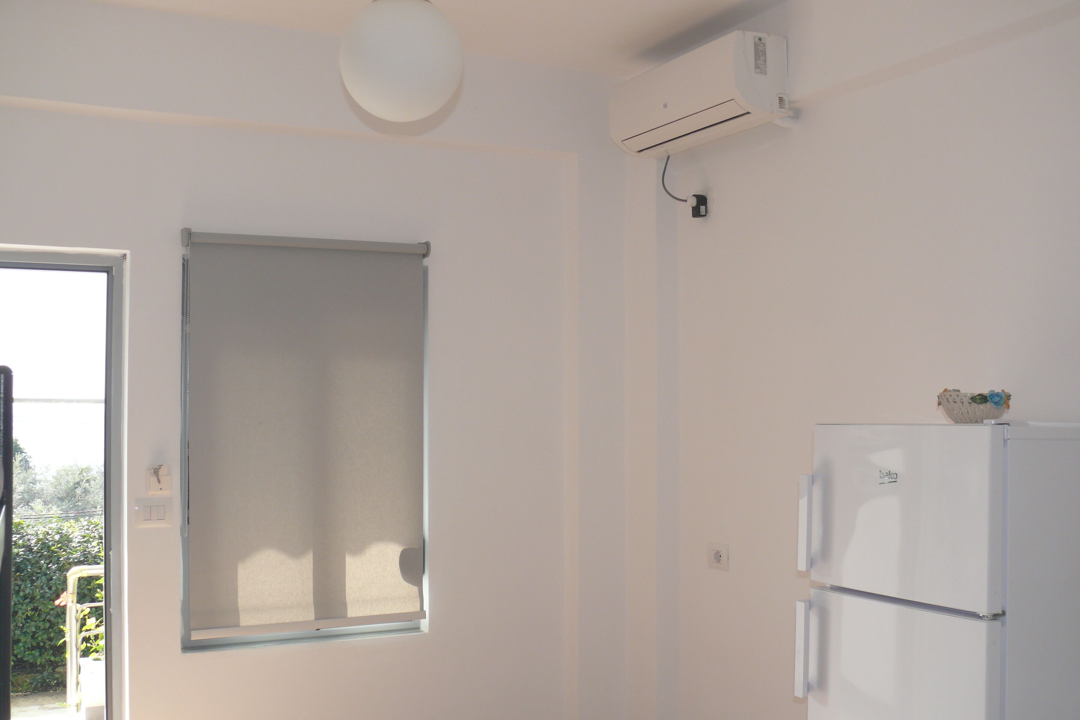 Una habitación con pared blanca, una ventana con persiana, un refrigerador blanco y un equipo de aire acondicionado.
