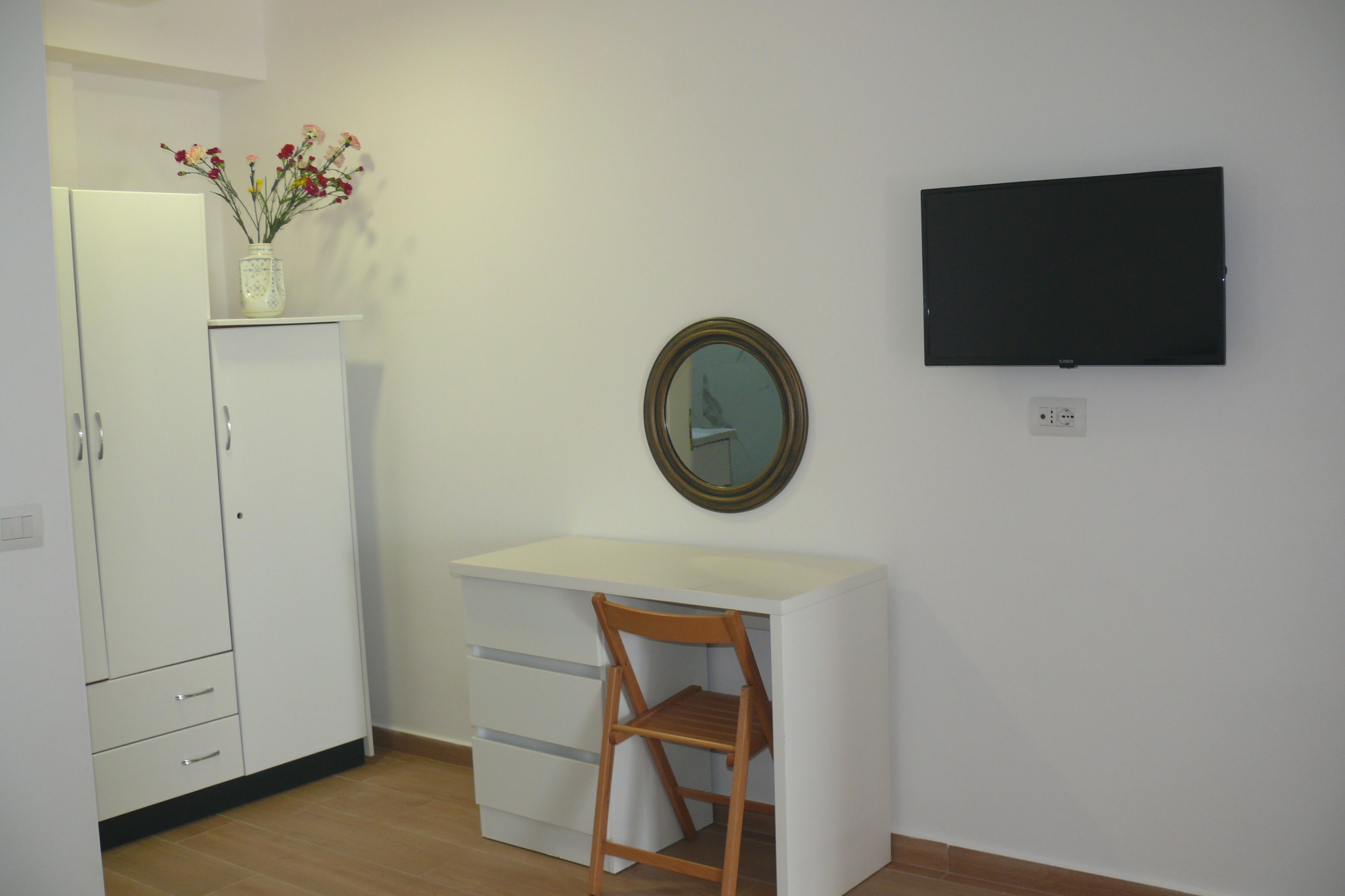 Quarto pequeno e limpo com escrivaninha branca, cadeira de madeira, guarda-roupa branco, espelho e televisão de tela plana presa à parede.