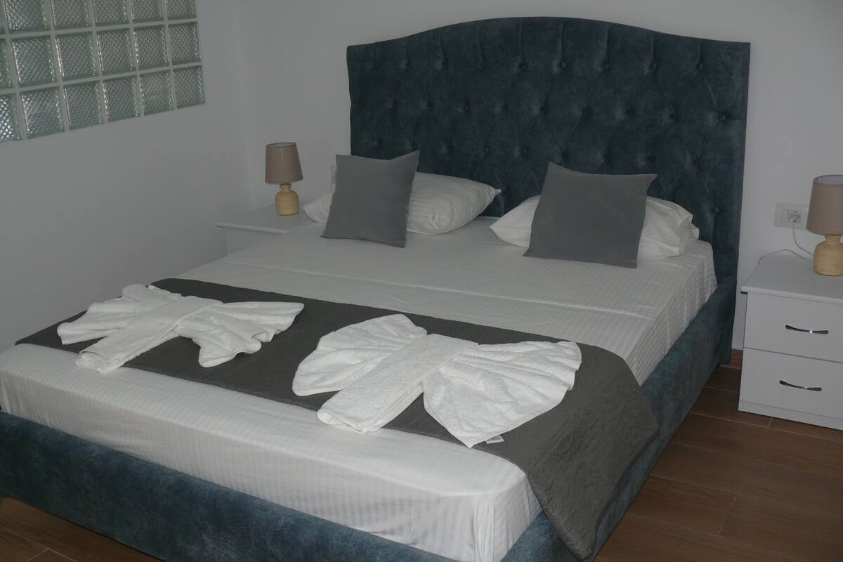 Una cama bien hecha con cabecera gris, ropa blanca de cama y almohada gris. Hay dos mesitas de noche con lámparas y un cajón pequeño.