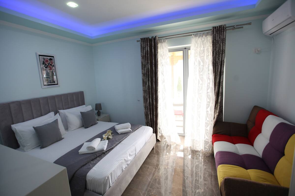 Habitación de hotel con cama, sofá, ventana con cortinas, un cuadro en la pared y un equipo de aire acondicionado.