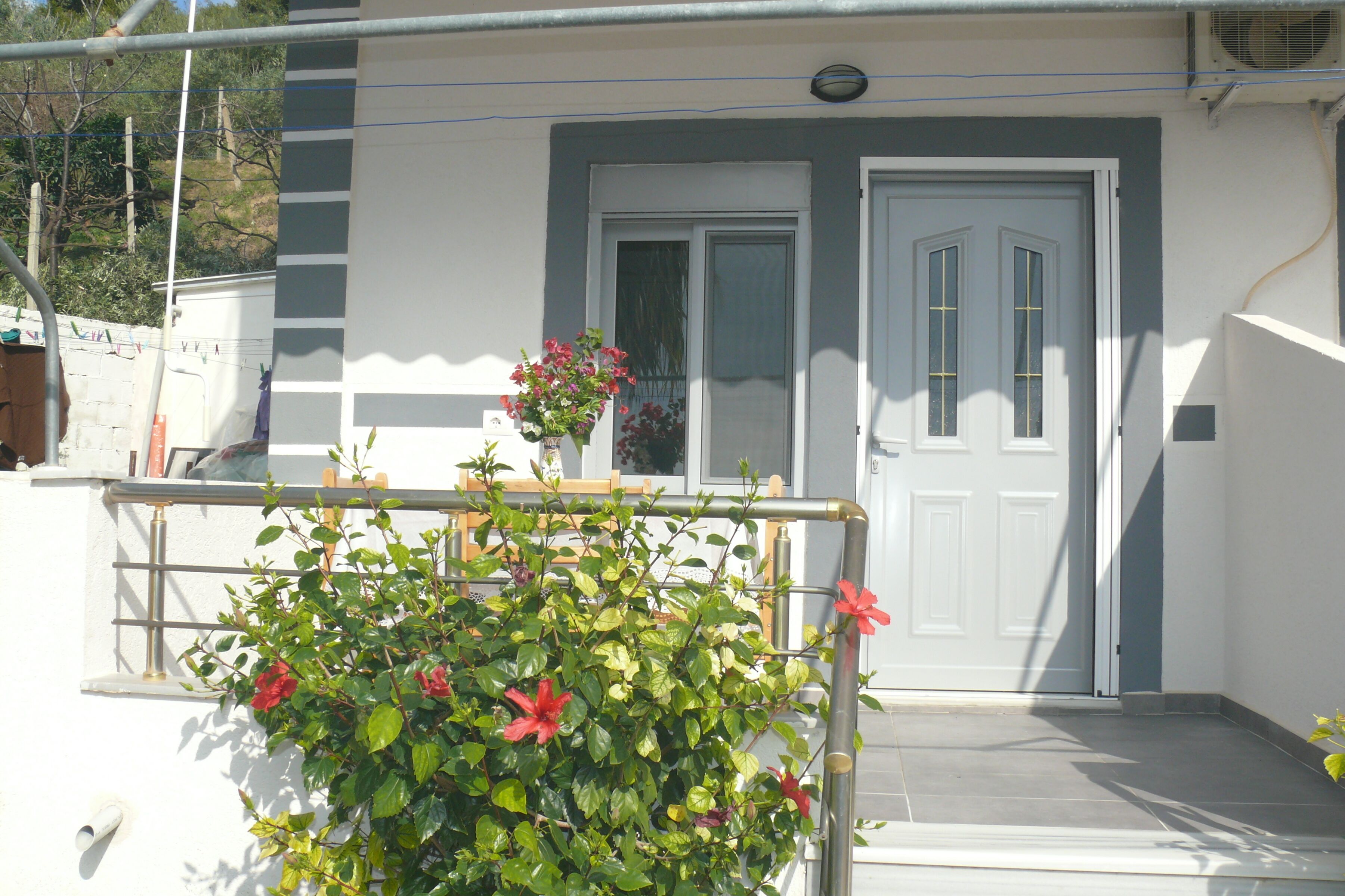 A white building with a grey door and a window. There is a metal railing in front of the building.