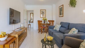 A living room with a TV, a dining table with chairs, a sofa, and a small table with a bowl of fruit and a bottle. - Avalon (Javea / Xàbia)