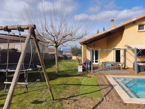 A backyard with a swing set, a small tree, a pool, and a patio area with outdoor furniture.