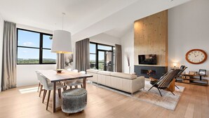 A modern living room with a dining area, large windows, a fireplace, and a TV mounted on the wall.
