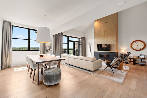 A modern living room with a dining area, large windows, a fireplace, and a TV mounted on the wall.