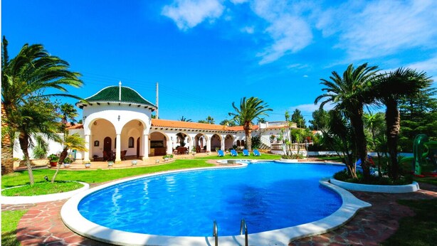 A swimming pool with a circular design, surrounded by palm trees and a building with a green roof and arches. - Villa Balneari Resort. Vacaciones en Familia y amigos Piscina Tenis Golf, Futbol (Rustical Balneario. Mont-roig del Camp)