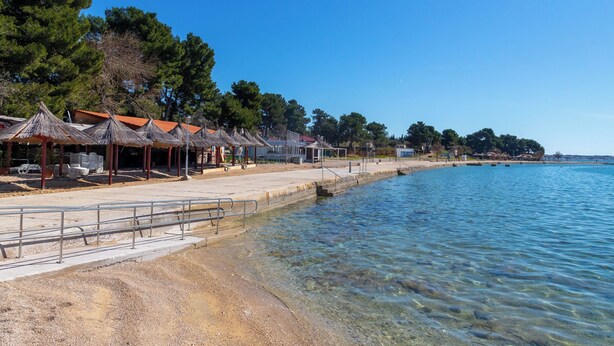 A beachside area with thatched-roof structures, clear shallow water, and a sandy beach. - Apartment Emina (Sv. Filip i Jakov)