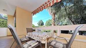 A balcony with a table and chairs, a striped awning, and a view of trees and a building. - Apartment Emina (Sv. Filip i Jakov)
