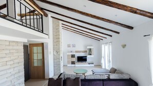A modern living room with a high ceiling, exposed beams, a stone wall, a sofa, a dining table with fruit, and a kitchen area in the background. - Apartment Marica (Supetarska Draga)