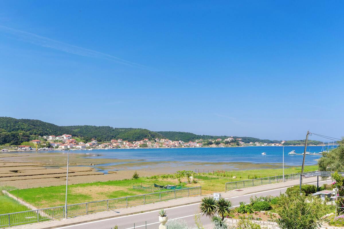 A coastal town with a clear view of the sea, a road with a fence, and residential buildings in the background.