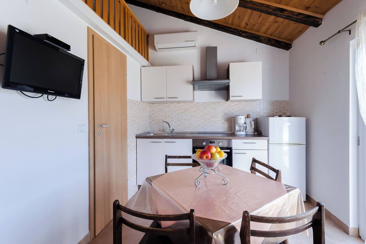 A compact kitchen with white cabinets, a built-in oven, and a refrigerator. There is a dining table with chairs and a bowl of fruit.