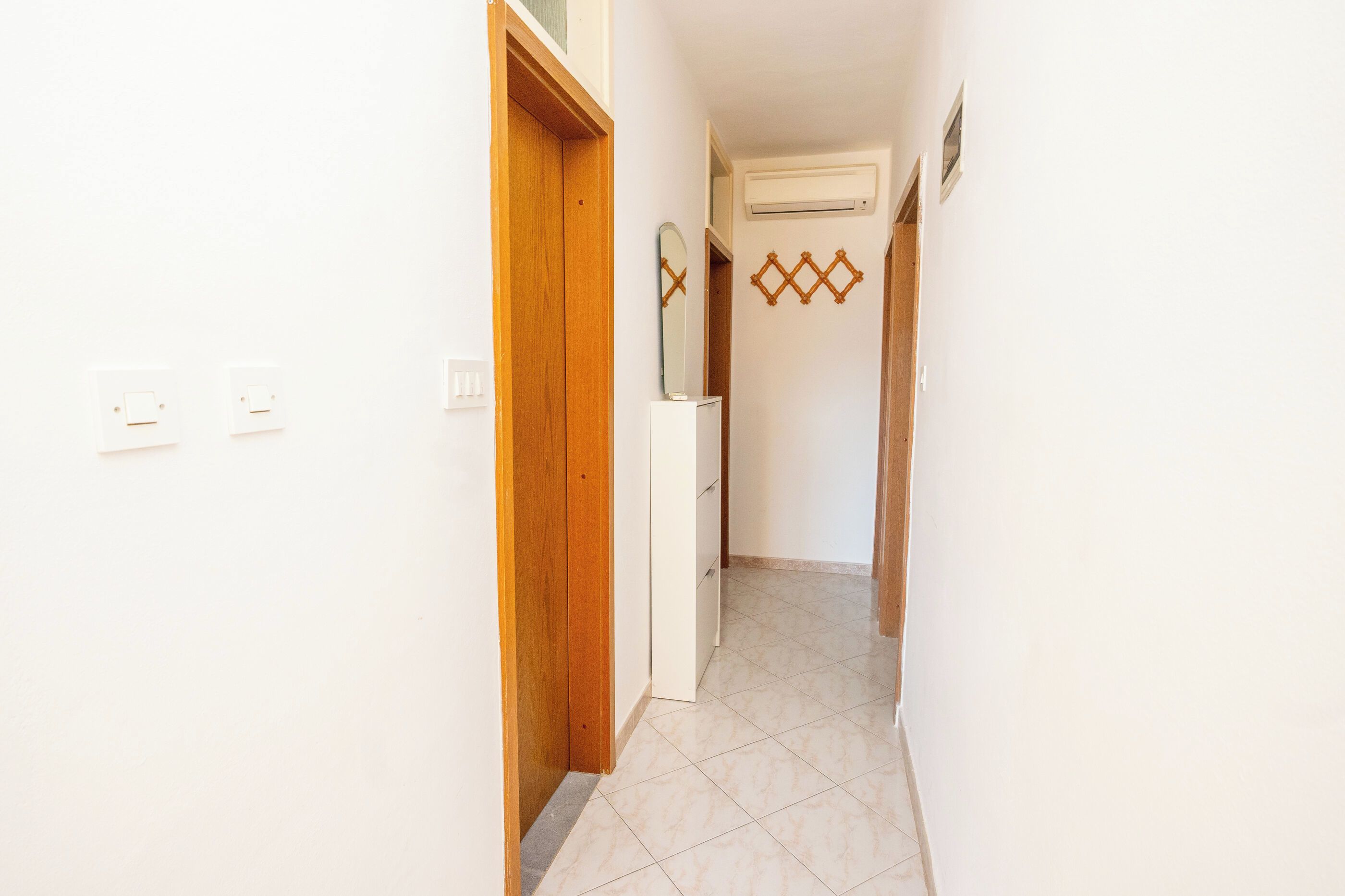 Un pasillo estrecho con una puerta de madera a la izquierda, una pared blanca con un interruptor de luz y un piso de baldosas.