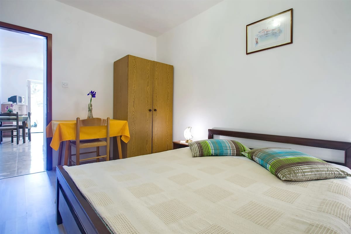 A bedroom with a bed, a wooden wardrobe, a table with a yellow tablecloth, and a chair.