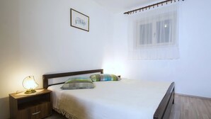 A bedroom with a bed, nightstand, lamp, and a window with sheer curtains. - Apartments Iris (Rabac)