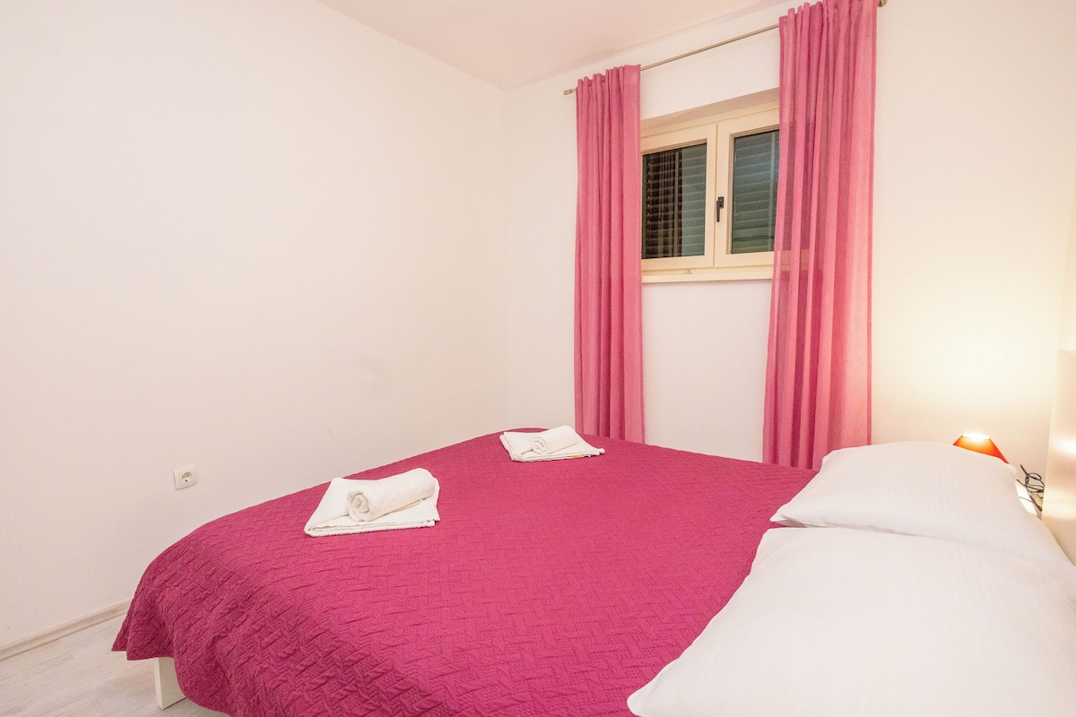 A single bed with a pink bedspread and white pillows, a window with pink curtains, and a bedside lamp.