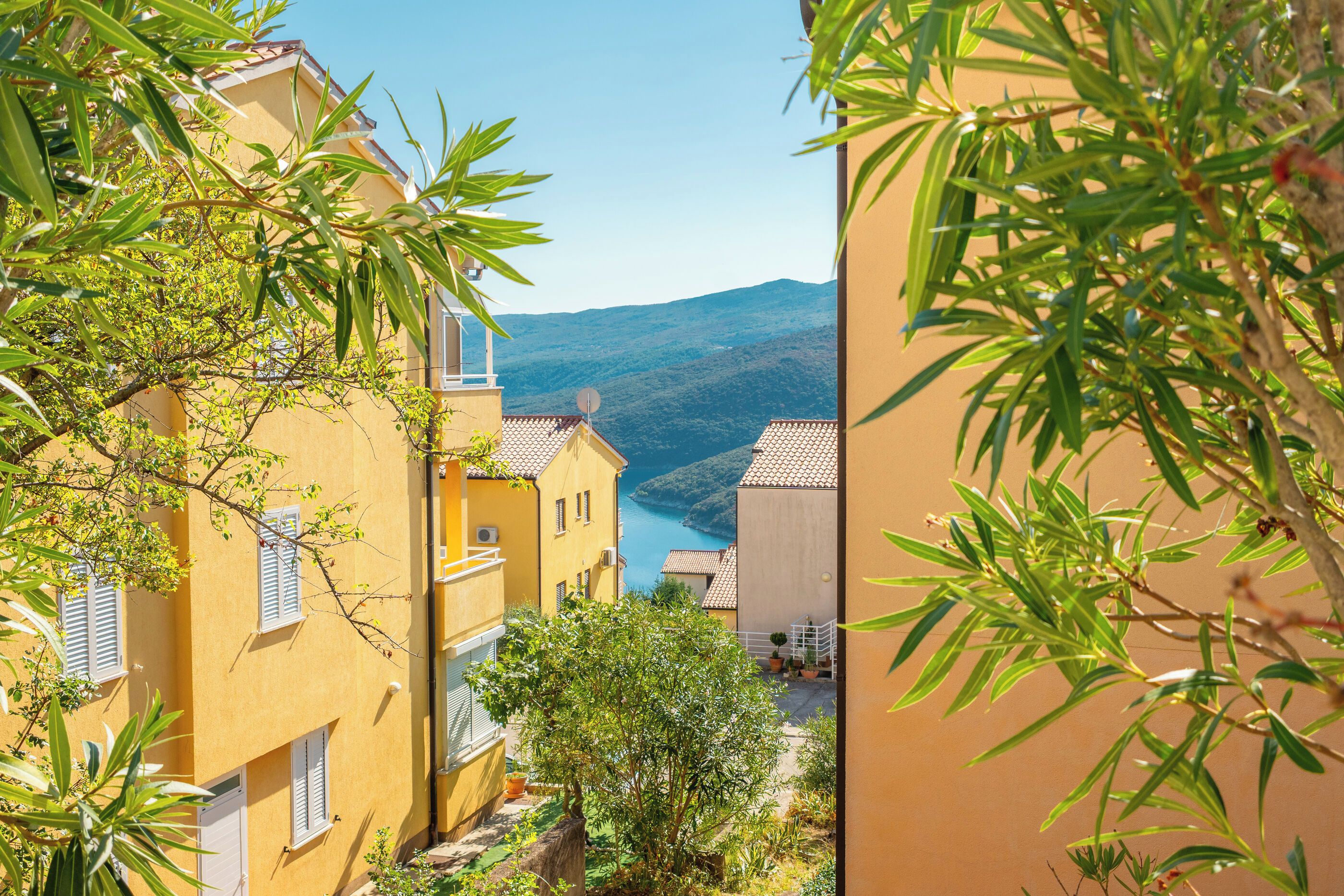 A narrow street with yellow buildings, a clear view of the sea, and a mountain in the distance.
