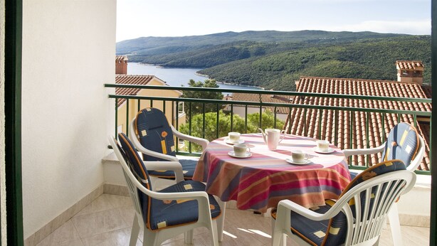 A balcony with a table and chairs, overlooking a scenic landscape with hills and a body of water. - Apartments Iris (Rabac)