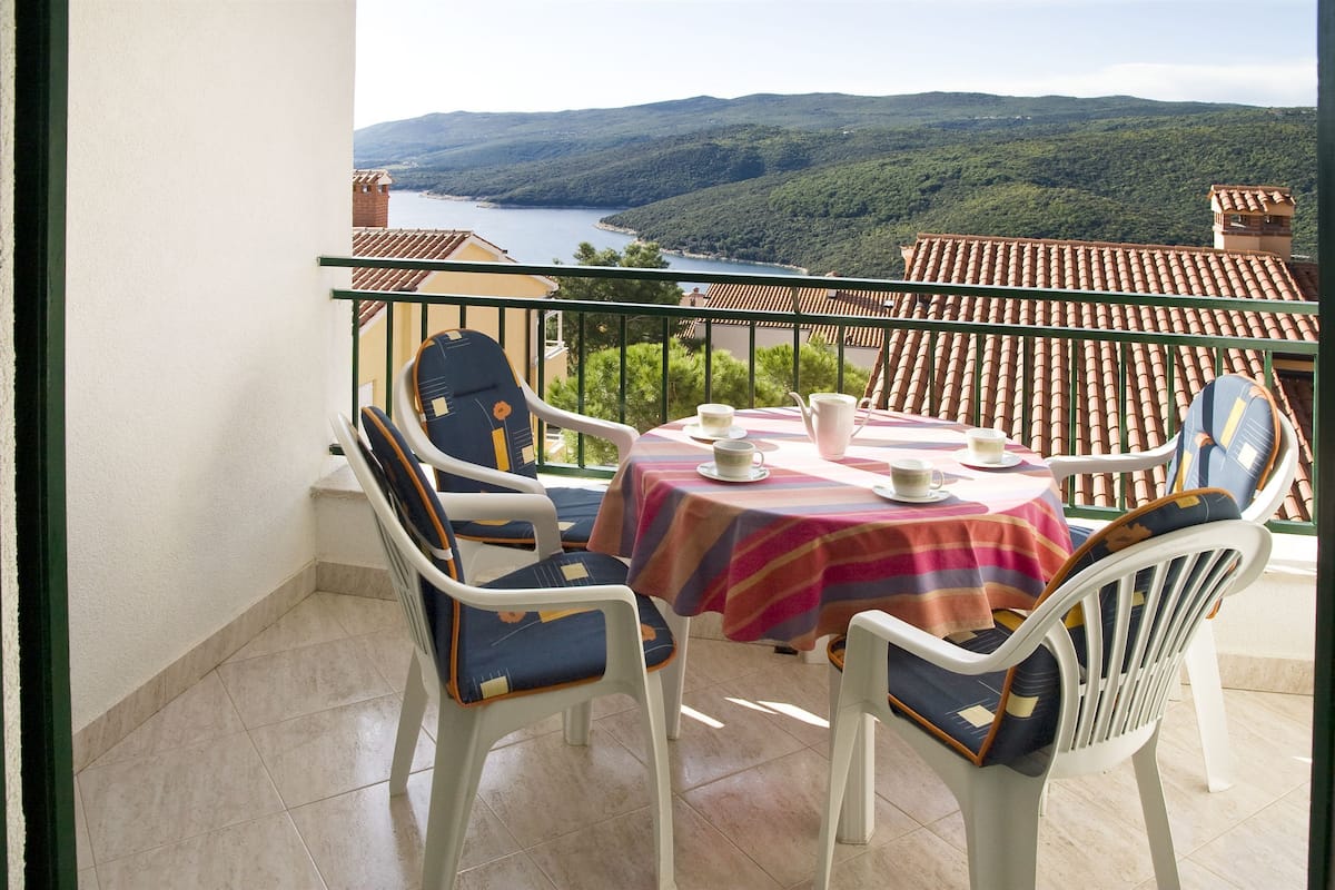 A balcony with a table and chairs, overlooking a scenic landscape with hills and a body of water.