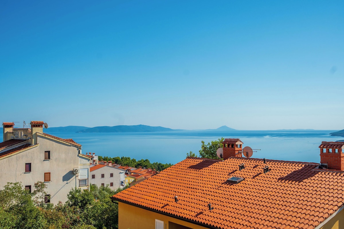 A coastal town with terracotta-roofed buildings, a clear blue sky, and a view of the sea and distant mountains.