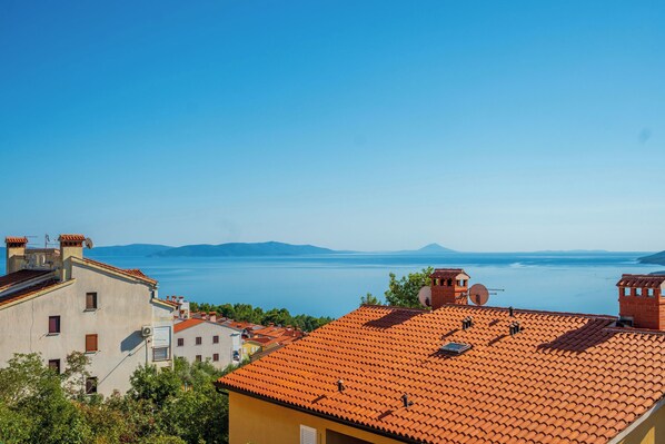 A coastal town with terracotta-roofed buildings, a clear blue sky, and a view of the sea and distant mountains.