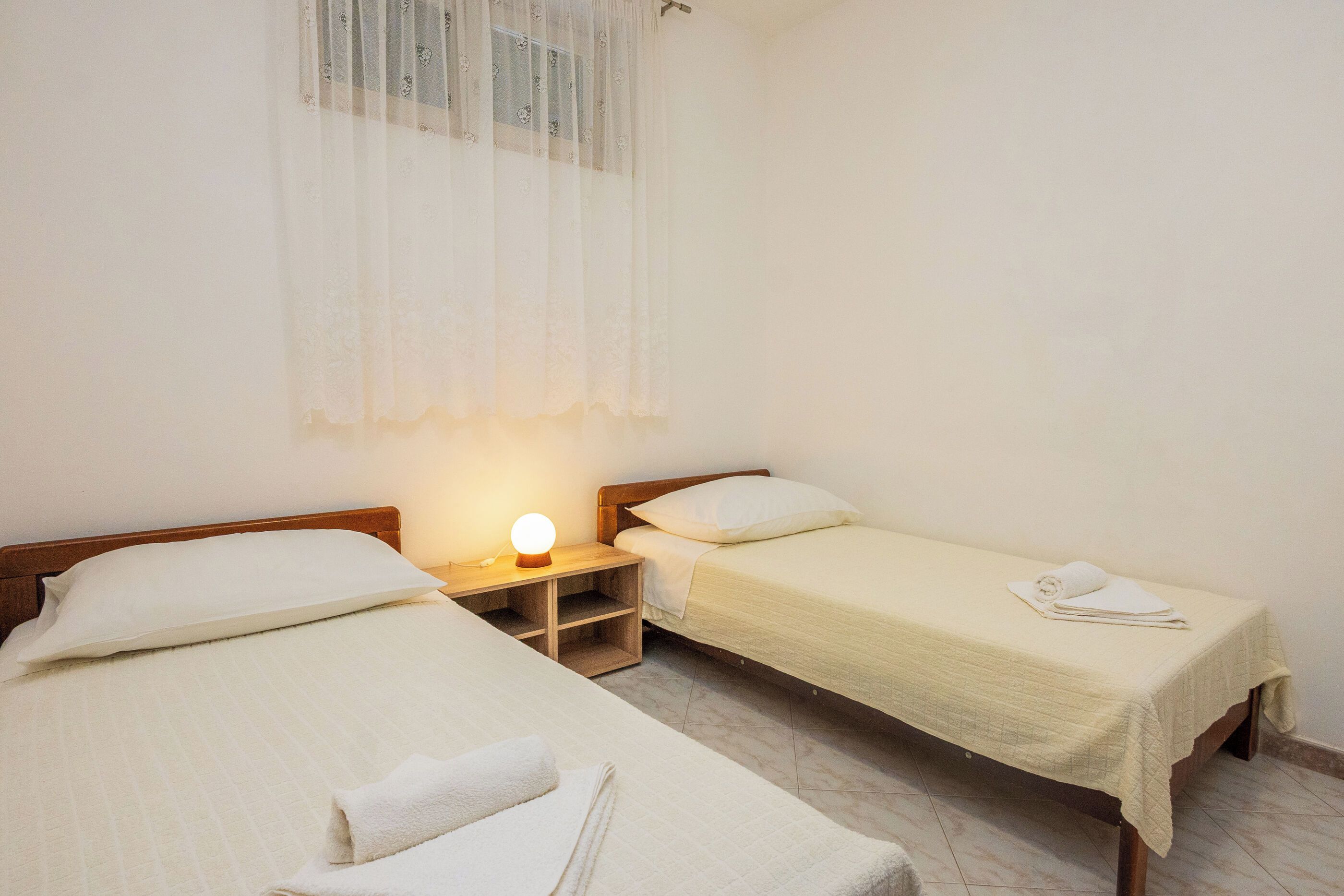 A room with two single beds, a bedside table with a lamp, and a window with sheer curtains.