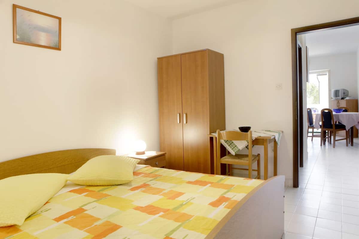 A bedroom with a bed, a wooden wardrobe, a desk with a chair, and a dining area visible through an open door.