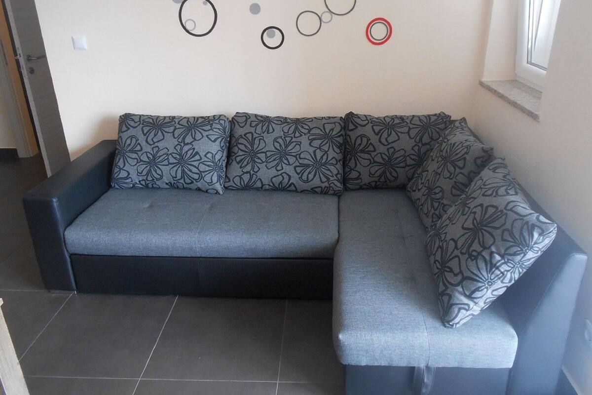 Un canapé d’angle gris avec des coussins à motifs floraux, dans une pièce dont le sol est recouvert de carreaux et un mur est orné de motifs circulaires abstraits.