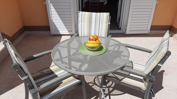 A round metal table with a woven top, surrounded by four metal chairs with cushioned seats. A bowl of fruit is placed on the table. - Apartments Pavelic (Povile)