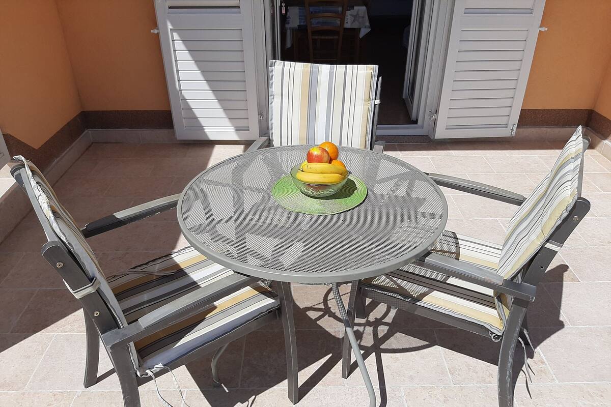 Une table ronde en métal, dont le plateau est recouvert d’un motif tissé, est entourée de quatre chaises en métal dotées d’assises rembourrées. Un saladier rempli de fruits est posé sur la table.