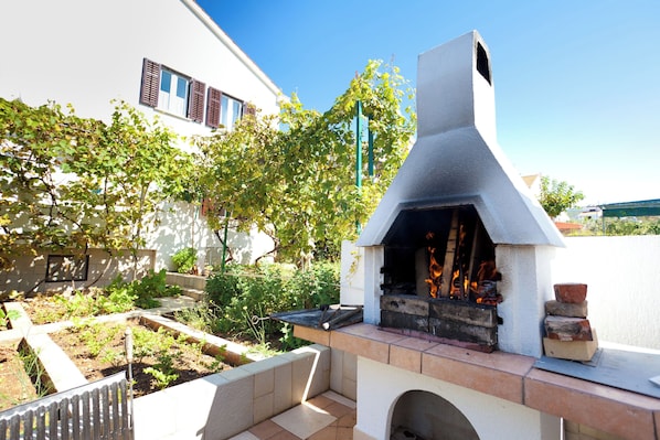 An outdoor barbecue area with a stone chimney, a wooden bench, and a garden with various plants.