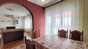 Ein Essbereich mit einem Tisch für vier Personen, Holzsesseln, einer Küche im Hintergrund und einem großen Fenster mit durchsichtigen Vorhängen.