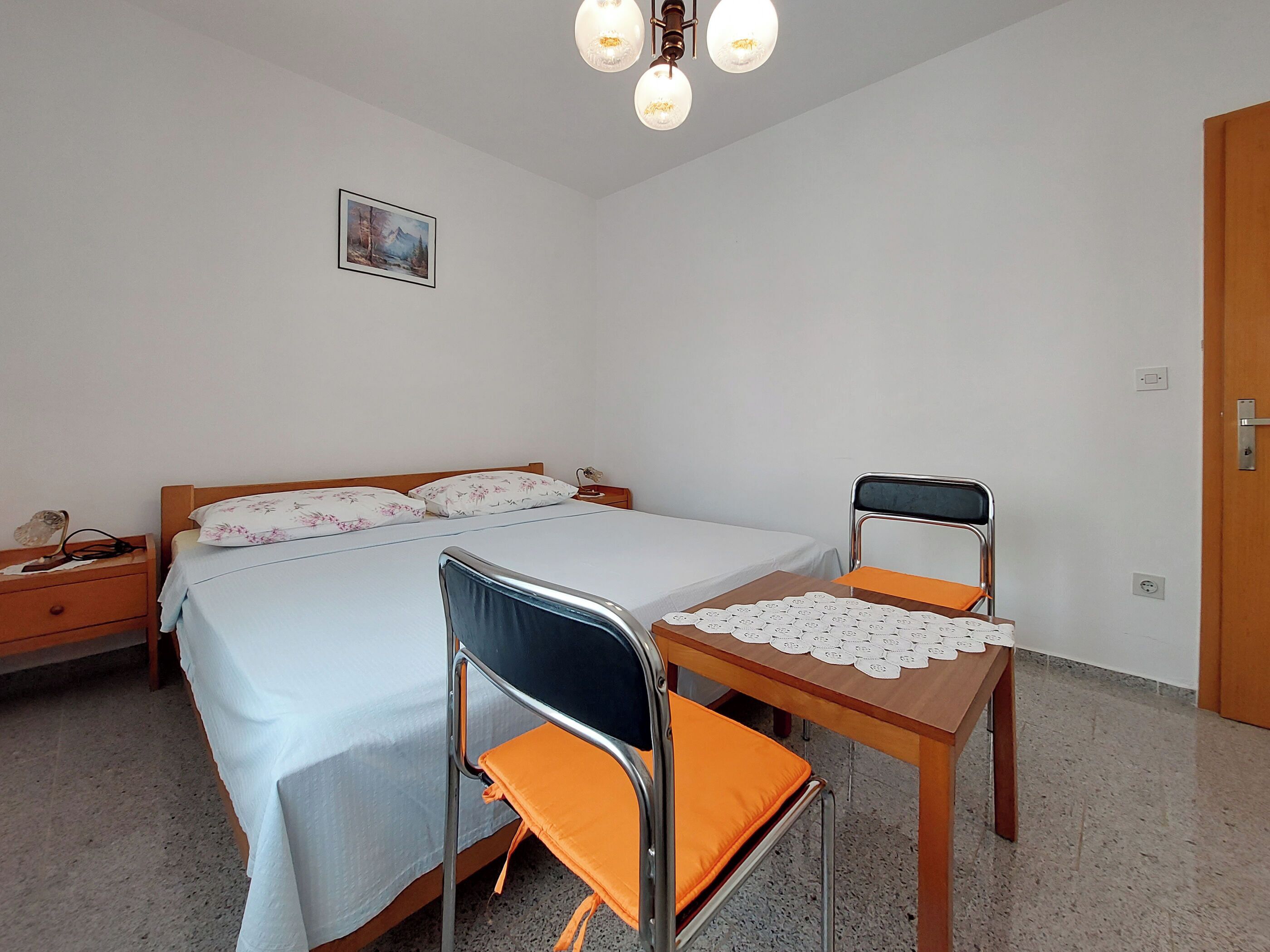 A bedroom with a bed, two chairs, a nightstand, a bedside table, a lamp, and a painting on the wall.