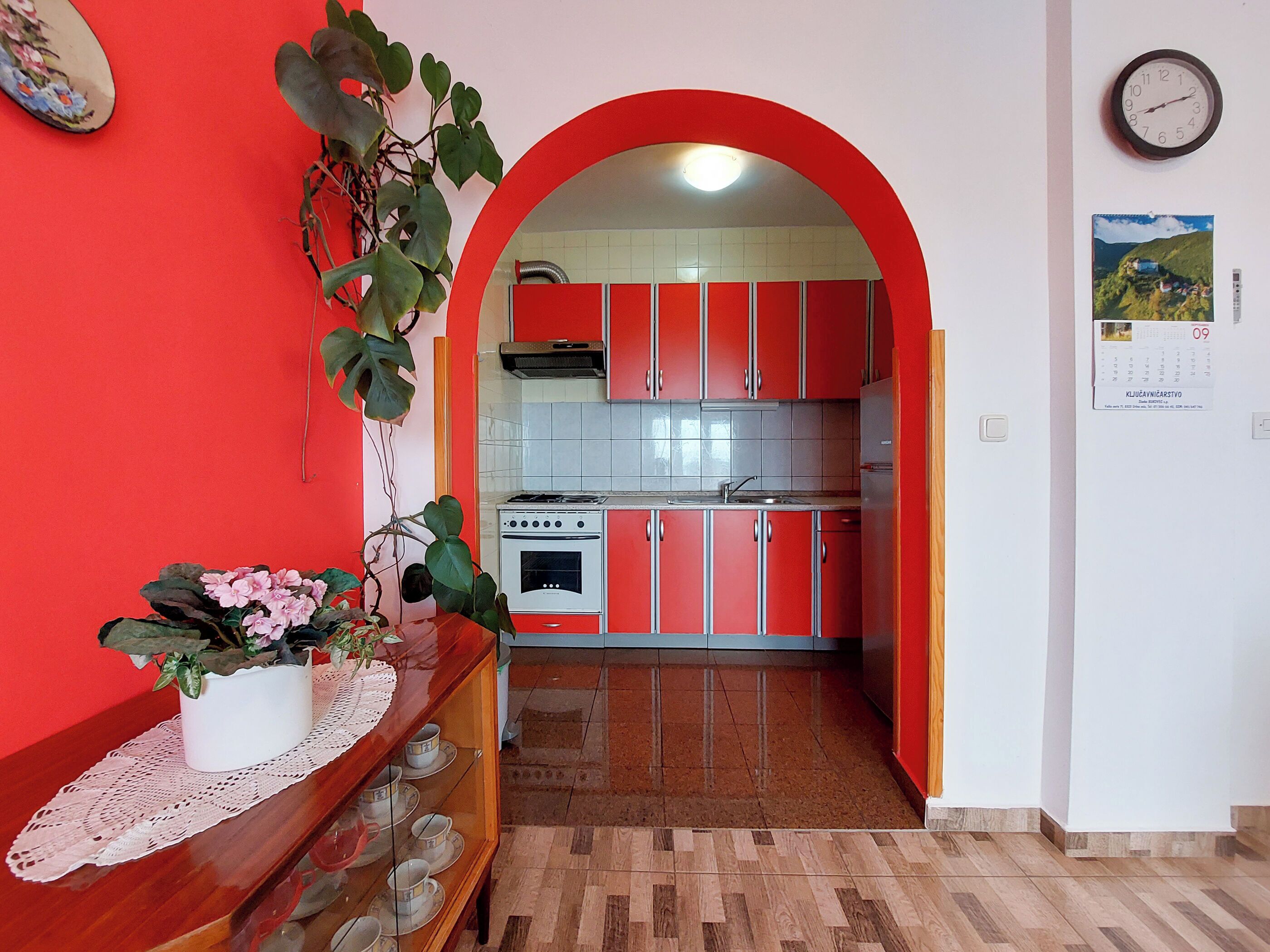 A kitchen with red cabinets, a stove, and a sink. There is a potted plant on a table in the foreground.