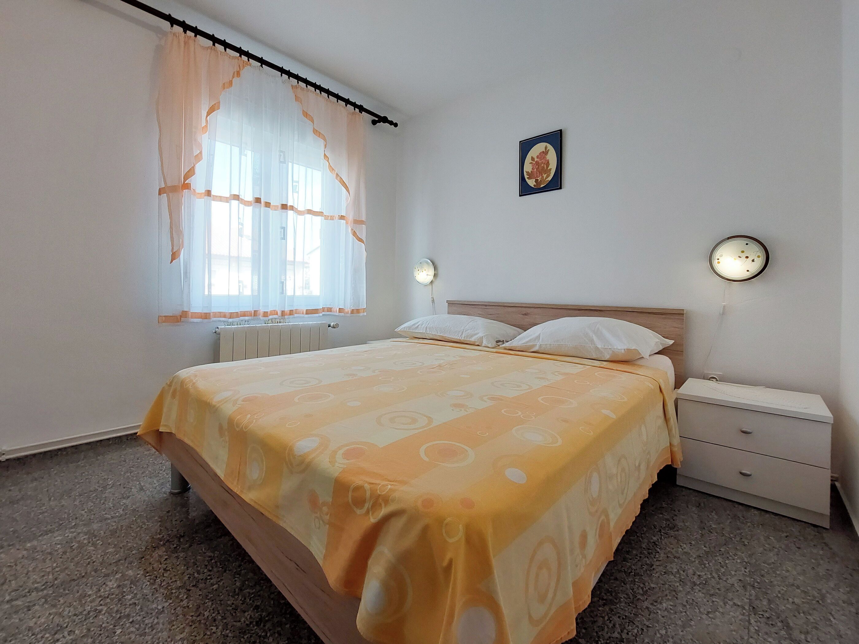 Un dormitorio con cama, mesita de noche, un reloj de pared y una ventana con cortinas traslúcidas.